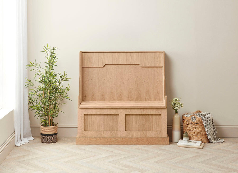 A wooden monks bench made from oak veneers, with a storage area beneath the seat, placed in a room with a plant and a basket.