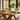 close up of solid pine tabletop showing natural grain, knots and warm finish with plates, glasses, and a vase with plants in a room with large windows showing a garden.