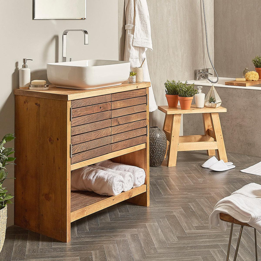 A freestanding solid wood bathroom vanity unit with two doors, a white sink on top, and a mirror above it. The vanity is placed in a bathroom setting with various decor items around it.